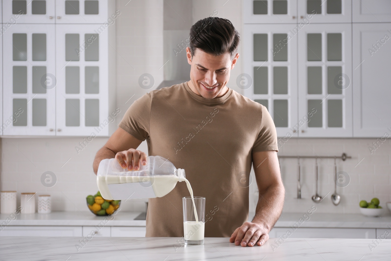 Photo of Man pouring milk from gallon bottle into glass at white marble table in kitchen