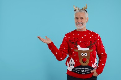Photo of Senior man in Christmas sweater and reindeer headband showing something on light blue background. Space for text