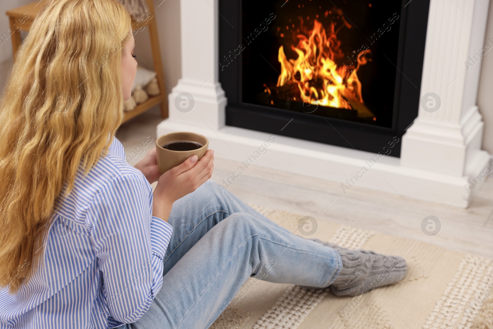 Photo of Young woman with cup of hot drink resting near fireplace at home