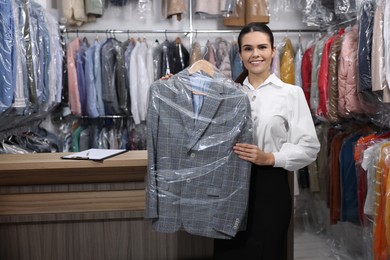 Dry-cleaning service. Happy worker holding hanger with jacket in plastic bag indoors, space for text