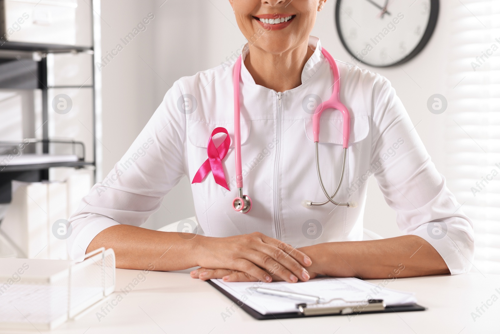 Photo of Doctor with stethoscope and pink ribbon at white desk indoors, closeup. Breast cancer awareness