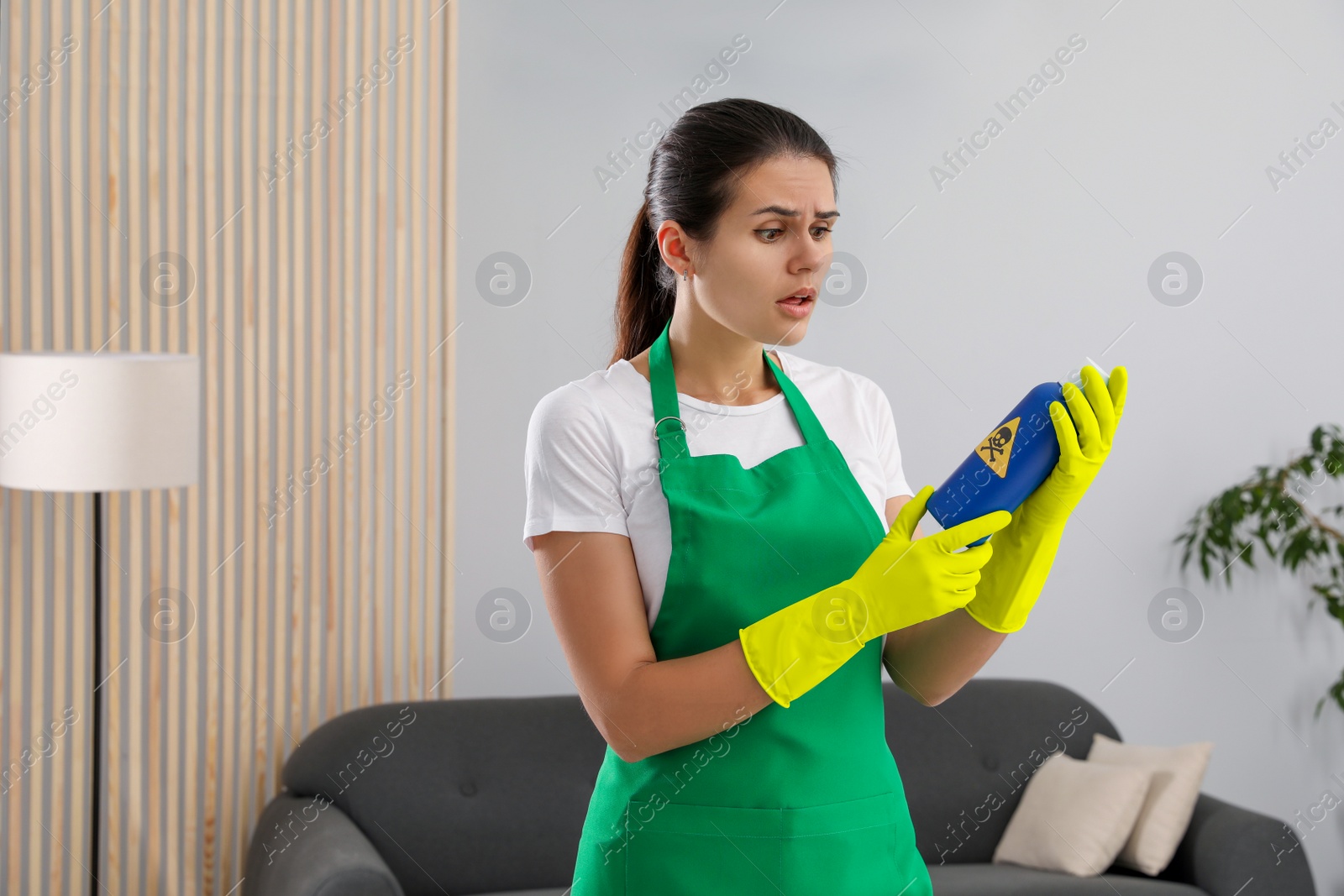 Photo of Woman looking at bottle of toxic household chemical with warning sign indoors