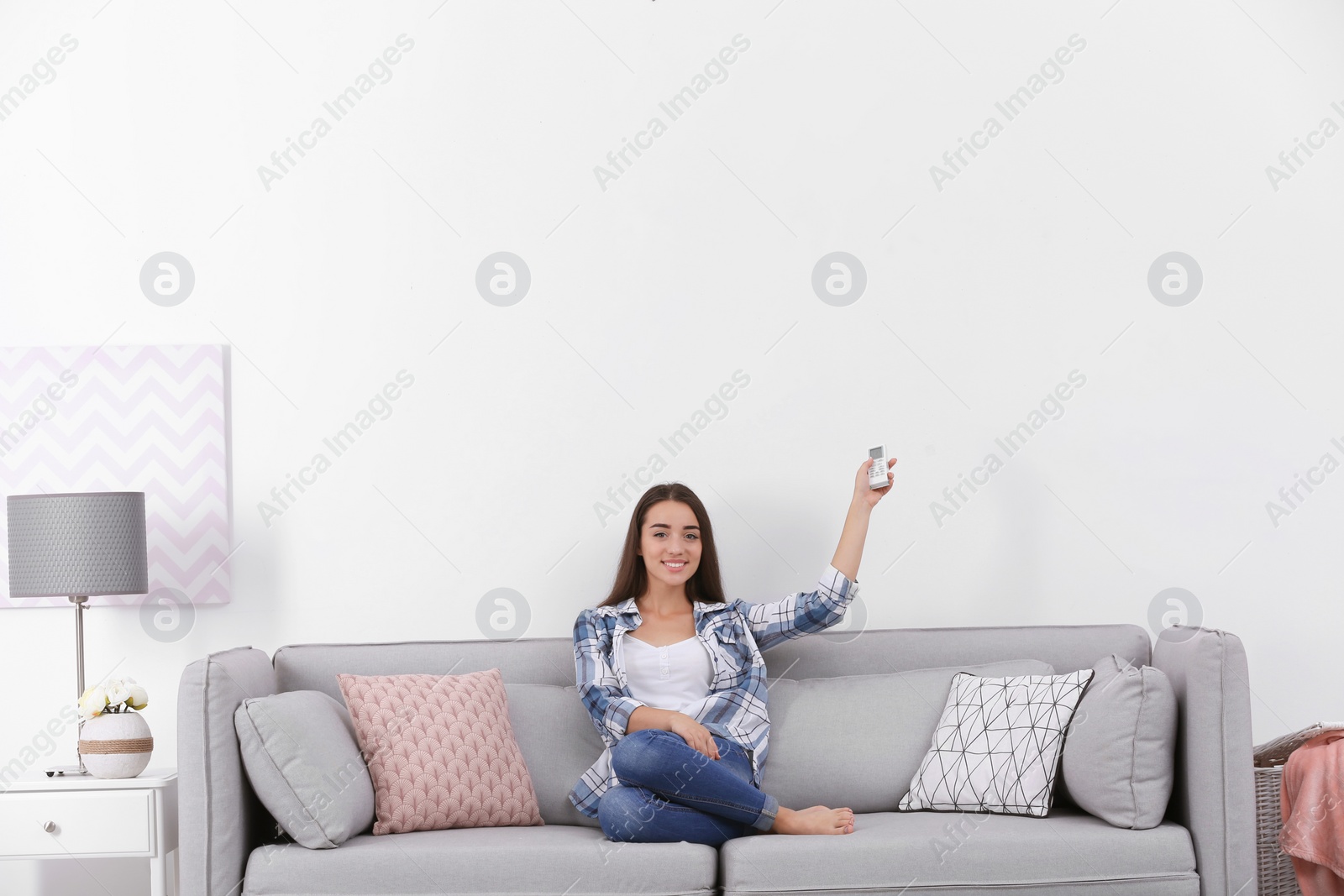 Photo of Young woman switching on air conditioner while sitting on sofa near white wall