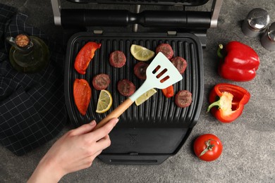 Photo of Woman cooking meat balls with bell peppers and lemon on electric grill at grey textured table, top view