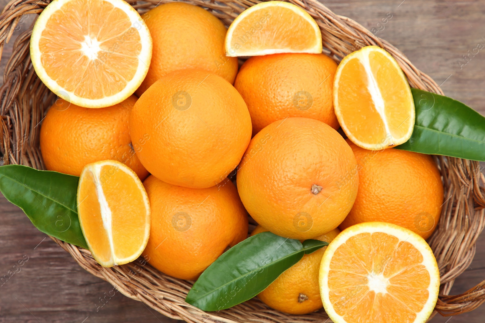 Photo of Wicker basket with ripe juicy oranges and green leaves on wooden table, top view