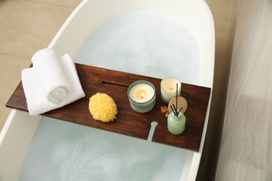 Photo of Wooden bath tray with candles, air freshener, towels and sponge on tub indoors, above view