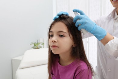 Photo of Doctor using lice treatment spray on little girl's hair indoors