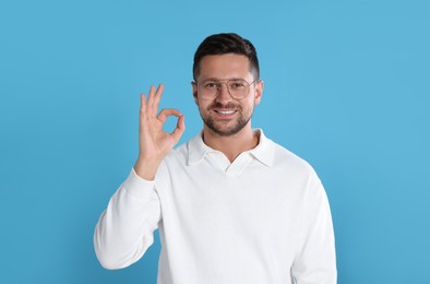 Portrait of happy man in stylish glasses showing ok gesture on light blue background