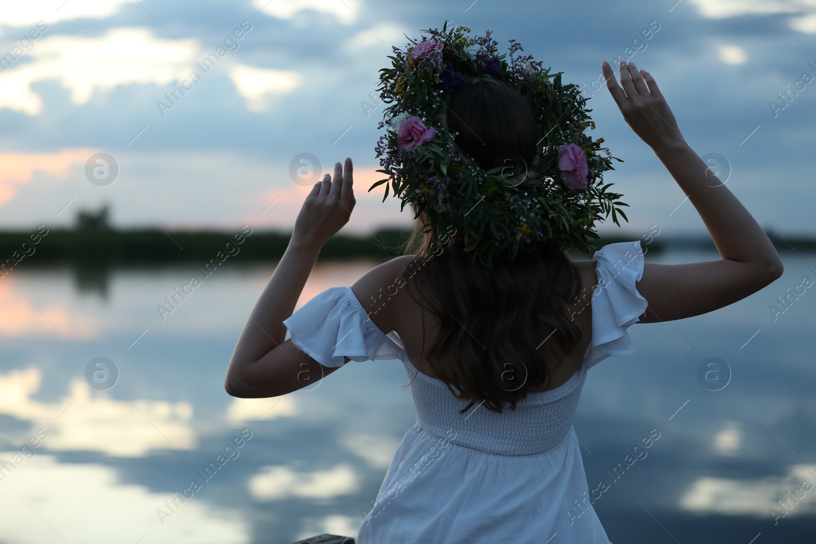 Photo of Young woman wearing wreath made of beautiful flowers near river in evening, back view