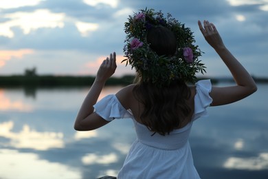 Young woman wearing wreath made of beautiful flowers near river in evening, back view