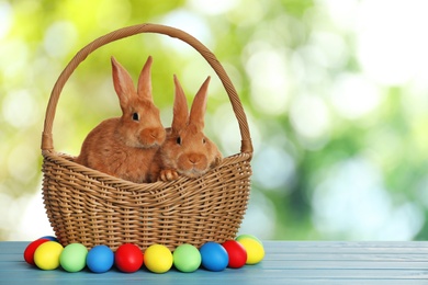 Adorable bunnies in wicker basket and Easter eggs on wooden surface outdoors, space for text