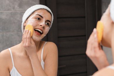 Photo of Young woman with headband washing her face using sponge near mirror in bathroom