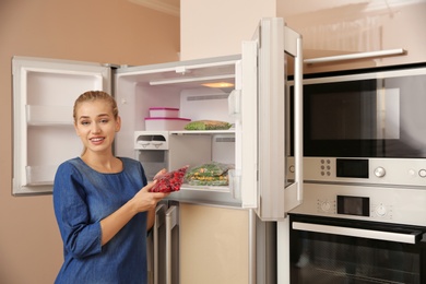 Photo of Young woman with frozen cherries near open refrigerator at home