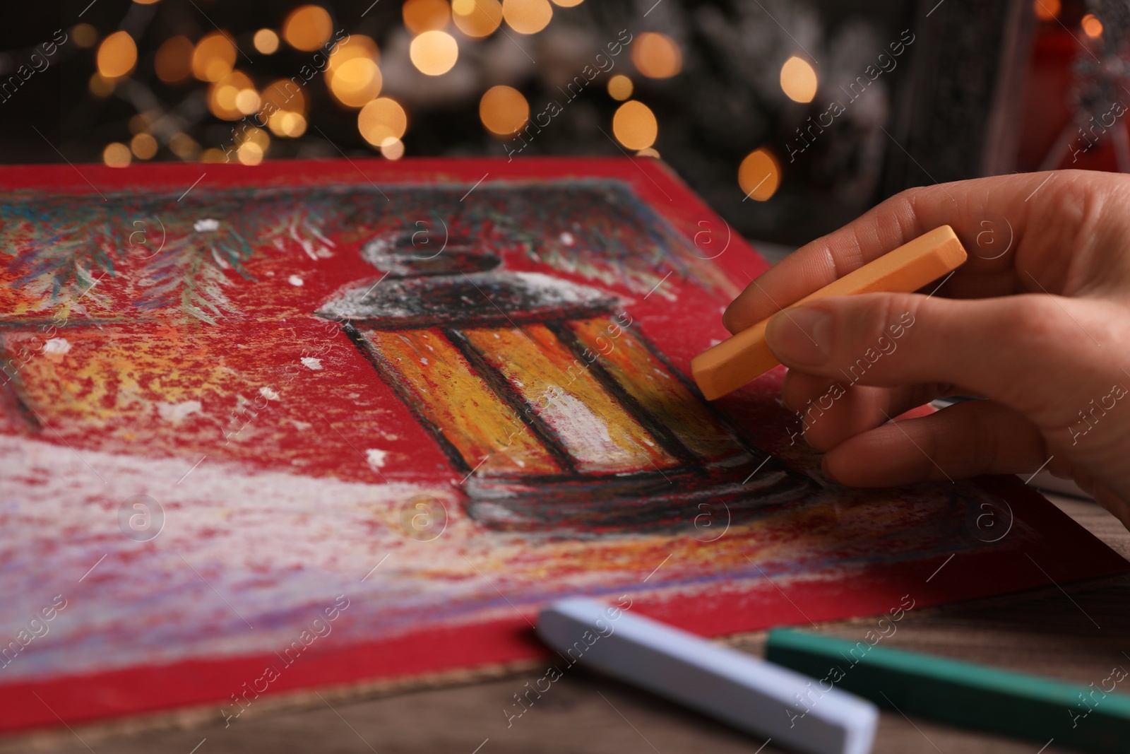 Photo of Woman drawing beautiful lantern with soft pastel at wooden table against blurred lights, closeup