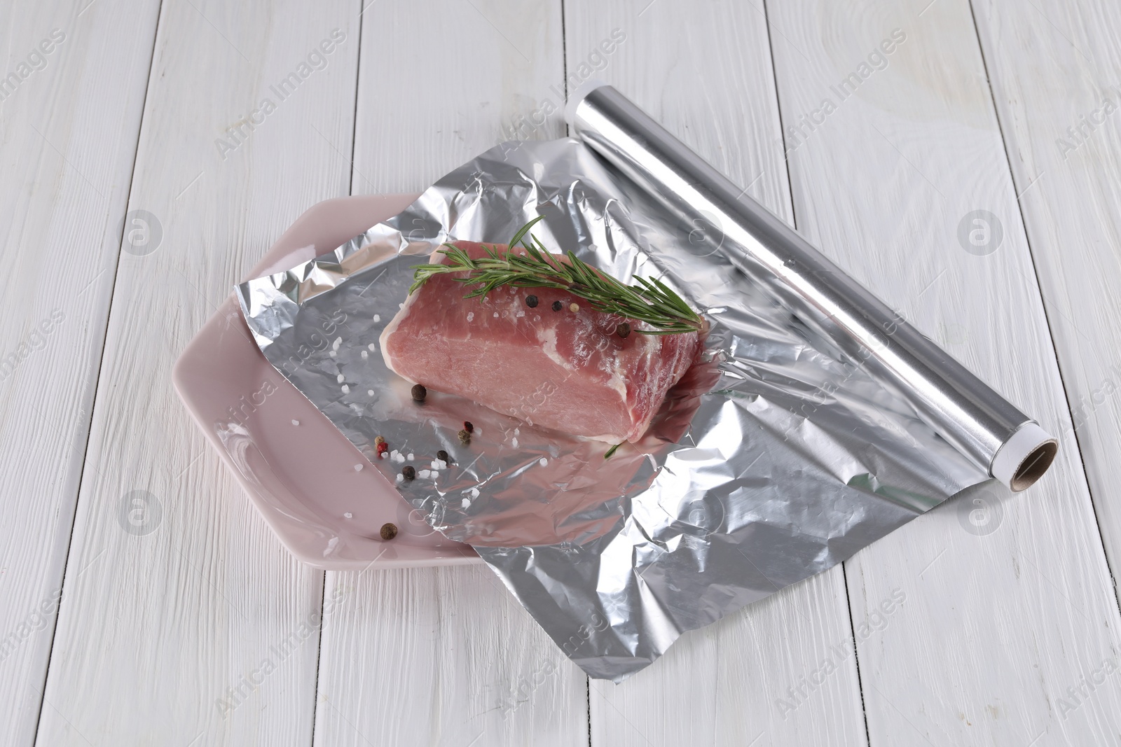 Photo of Aluminum foil with raw meat, rosemary and spices on white wooden table