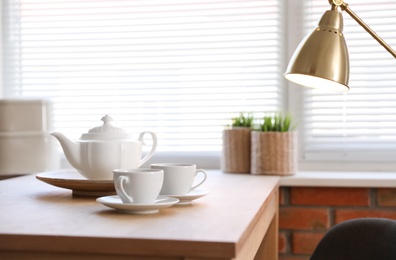 Photo of Tea set on table near window with blinds indoors