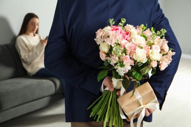 Man hiding bouquet of flowers and present for his beloved woman indoors, closeup