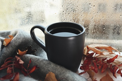 Cup of hot drink and autumn leaves near window on rainy day. Cozy atmosphere