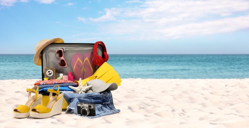 Image of Suitcase with beach accessories on sand near ocean, space for text