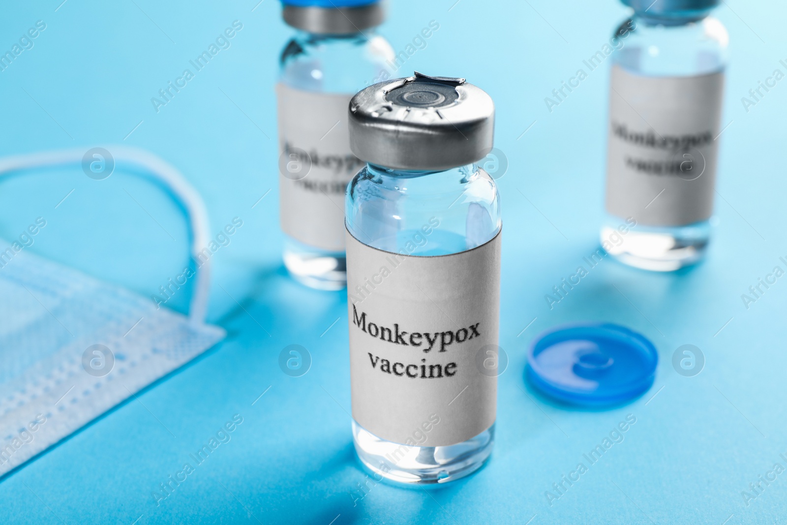 Photo of Monkeypox vaccine in glass vials and medical mask on light blue background