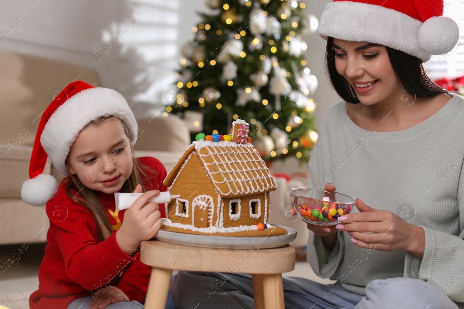 Photo of Mother and daughter decorating gingerbread house at table indoors