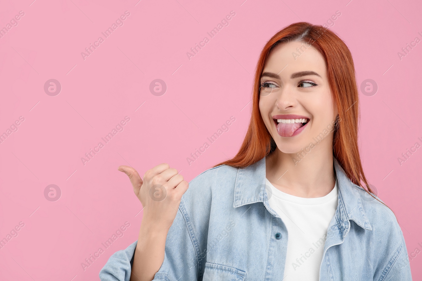 Photo of Happy woman showing her tongue and pointing at something on pink background. Space for text