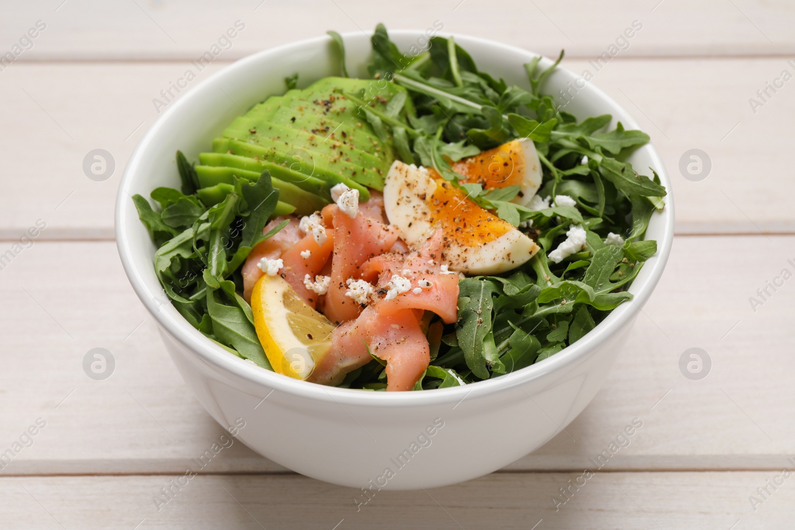 Photo of Delicious salad with boiled egg, salmon and avocado in bowl on white wooden table, closeup