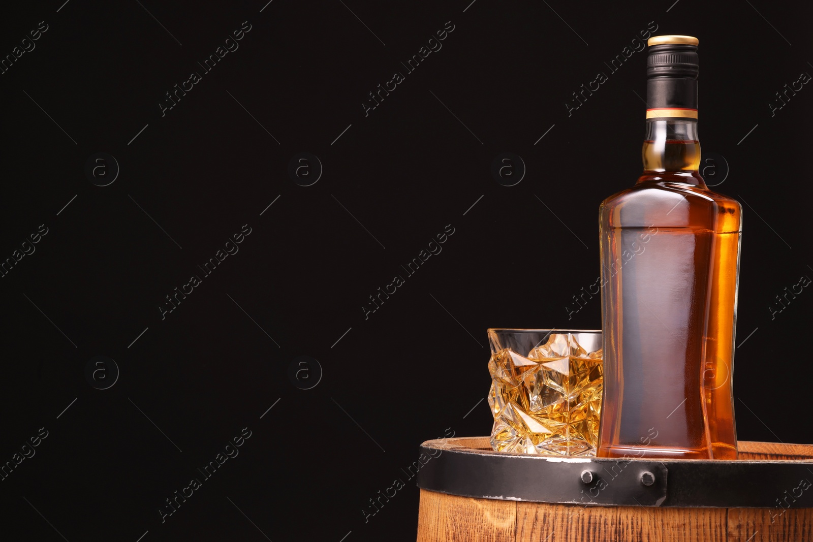 Photo of Whiskey in glass and bottle on wooden barrel against black background, space for text