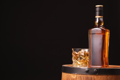 Whiskey in glass and bottle on wooden barrel against black background, space for text