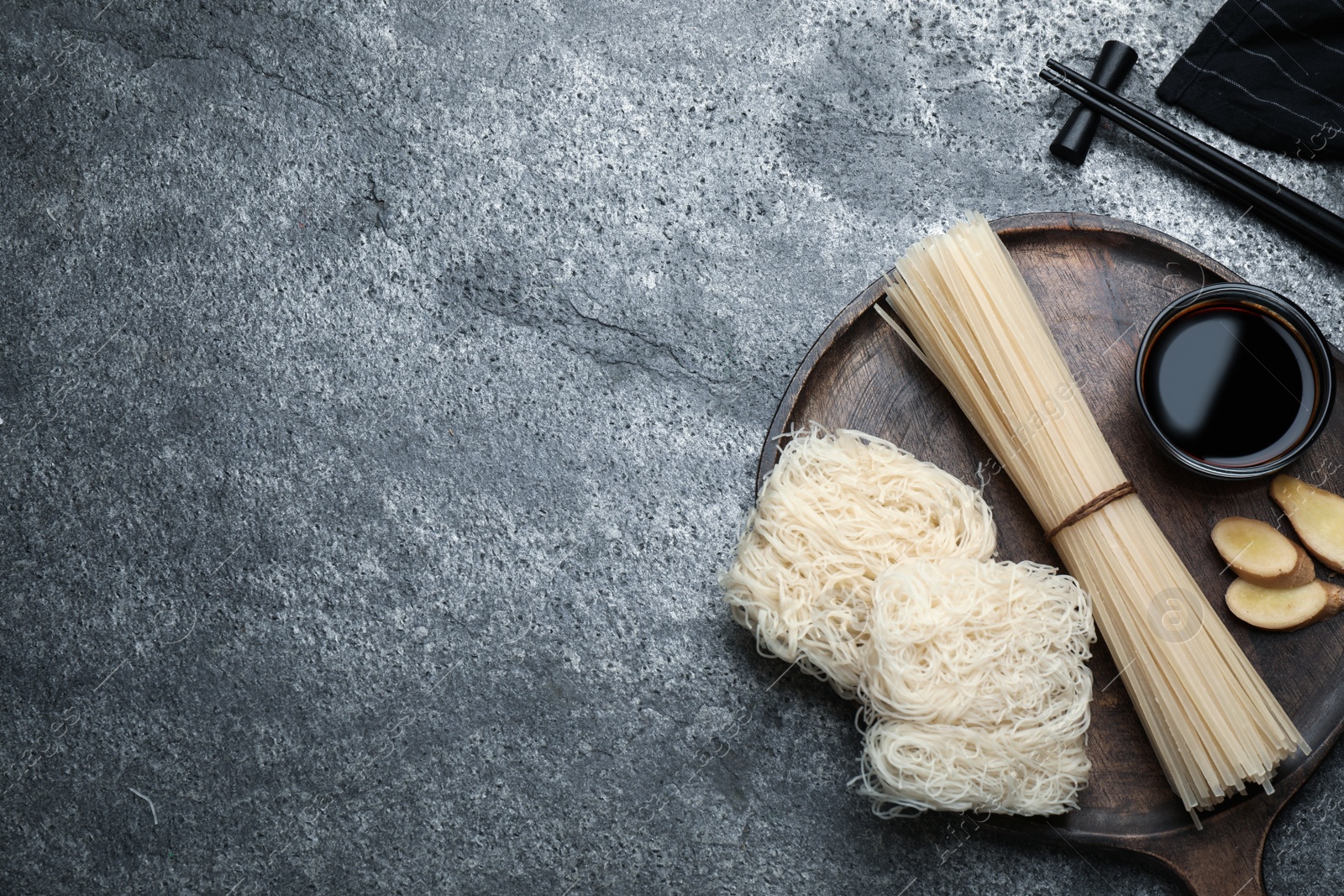 Photo of Different uncooked rice noodles, soy sauce, cut ginger and chopsticks on grey table, flat lay. Space for text