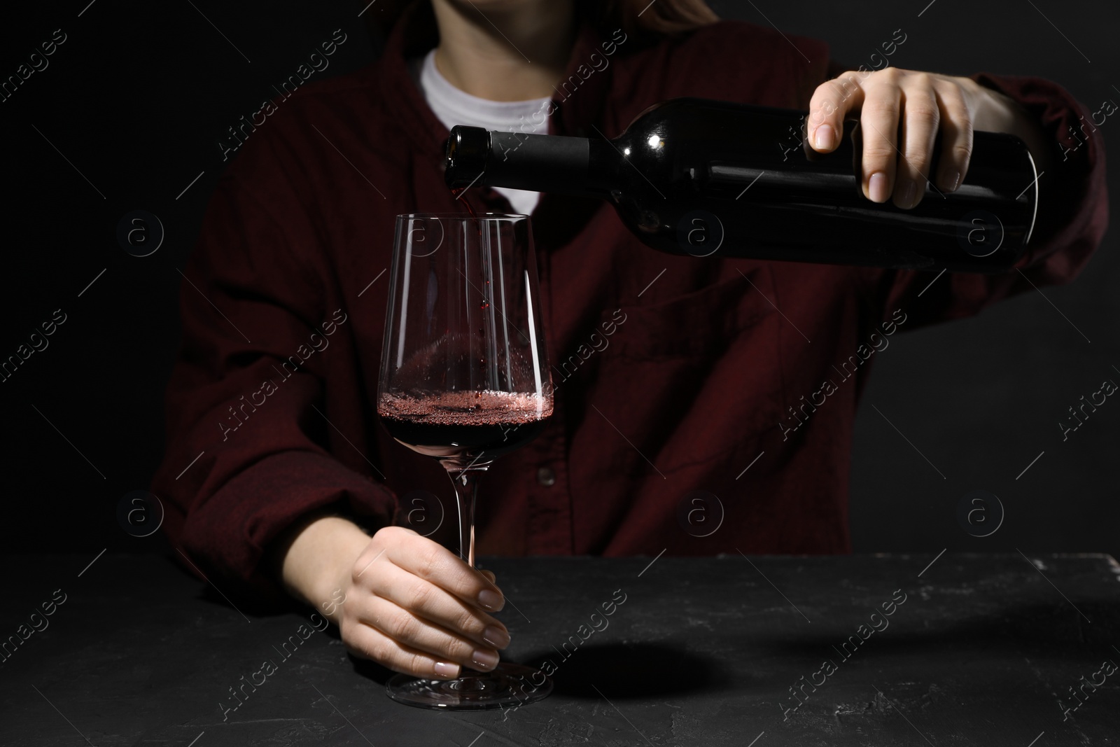 Photo of Alcohol addiction. Woman pouring red wine from bottle into glass at dark textured table, closeup