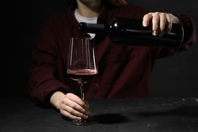 Photo of Alcohol addiction. Woman pouring red wine from bottle into glass at dark textured table, closeup