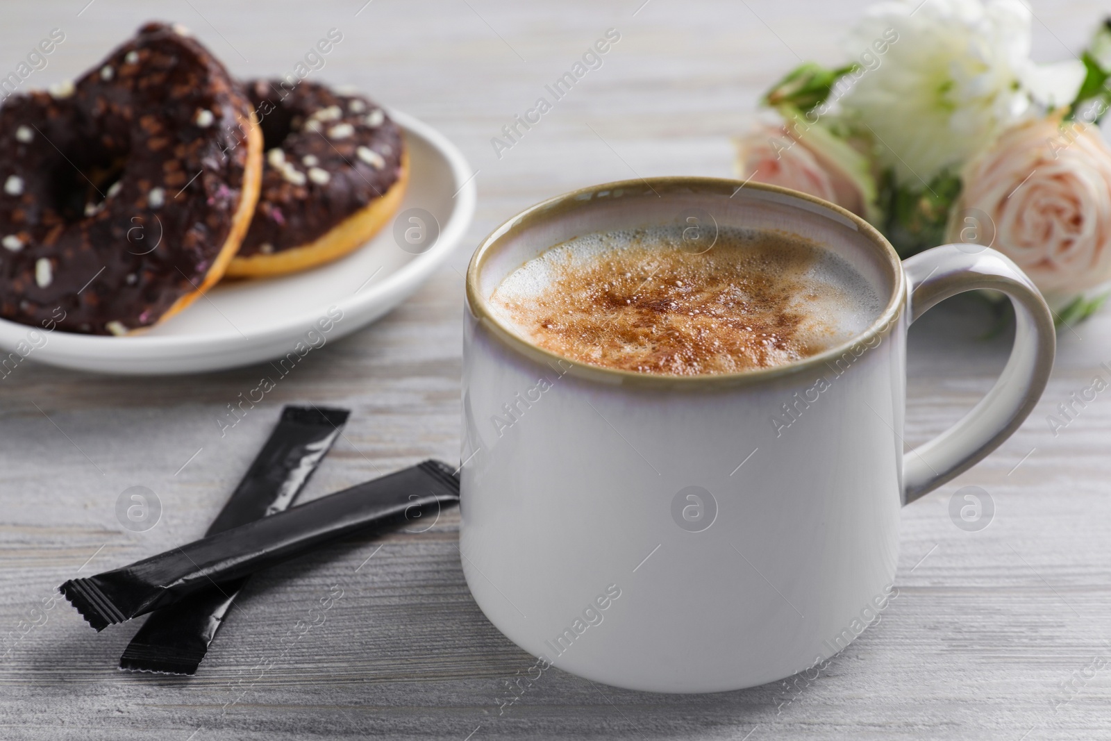 Photo of Delicious coffee in cup, sugar packets, doughnuts and flowers on white wooden table