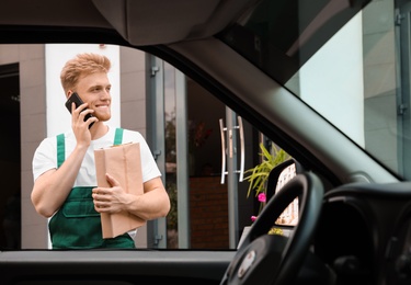 Photo of Young courier talking on phone near delivery car outdoors, view from inside