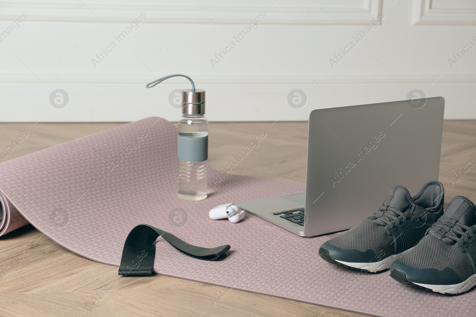 Photo of Exercise mat, laptop, bottle of water, wireless earphones, fitness elastic band and shoes on wooden floor indoors