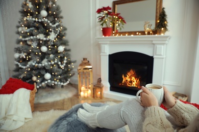 Photo of Woman with hot drink resting near fireplace in cozy room decorated for Christmas, closeup