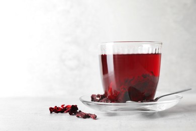 Photo of Aromatic hibiscus tea in glass, dried roselle calyces and spoon on light table, space for text