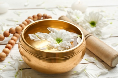 Photo of Golden singing bowl with flower, mallet and beads on white wooden table, closeup. Sound healing