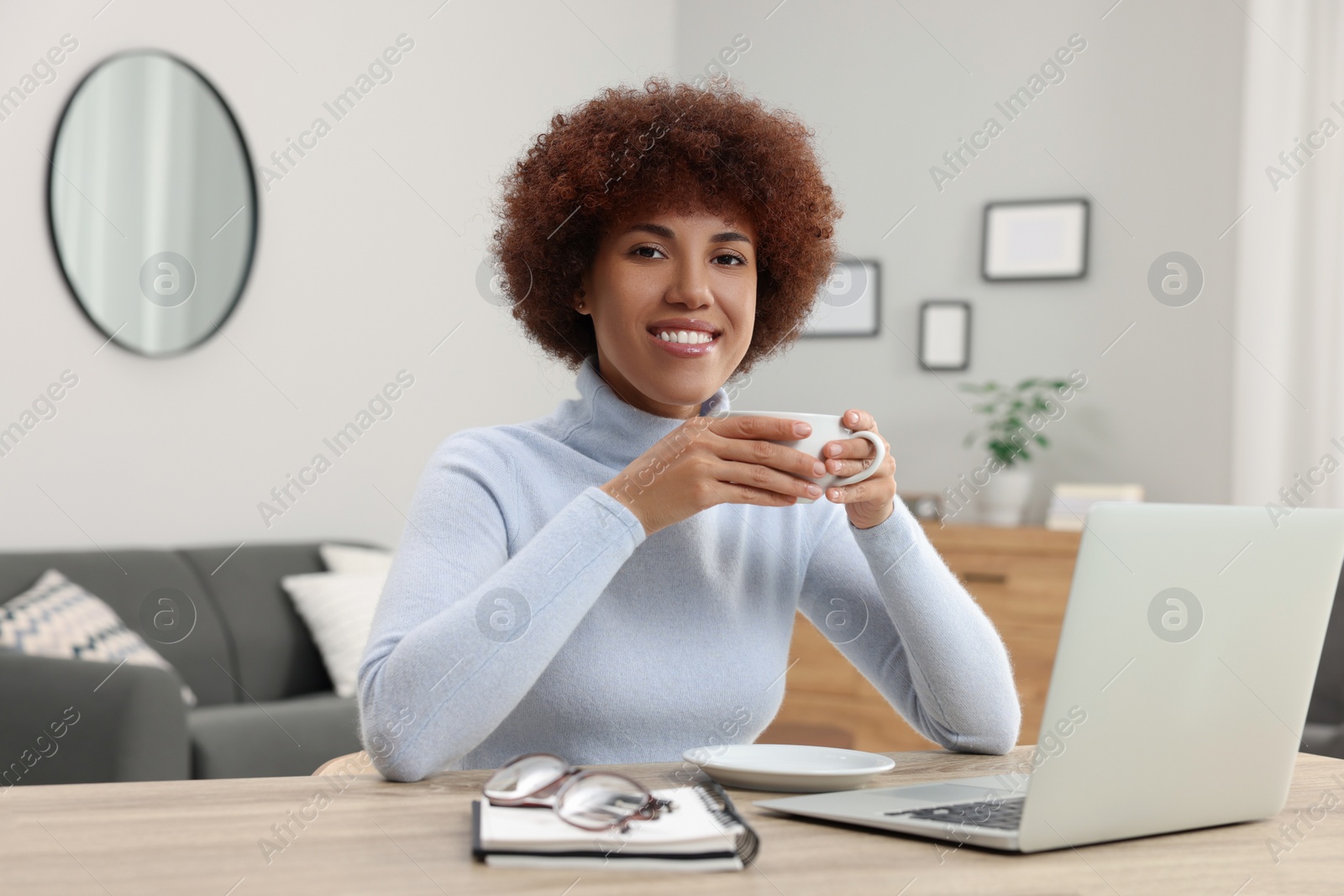 Photo of Beautiful young woman using laptop and drinking coffee at wooden desk in room