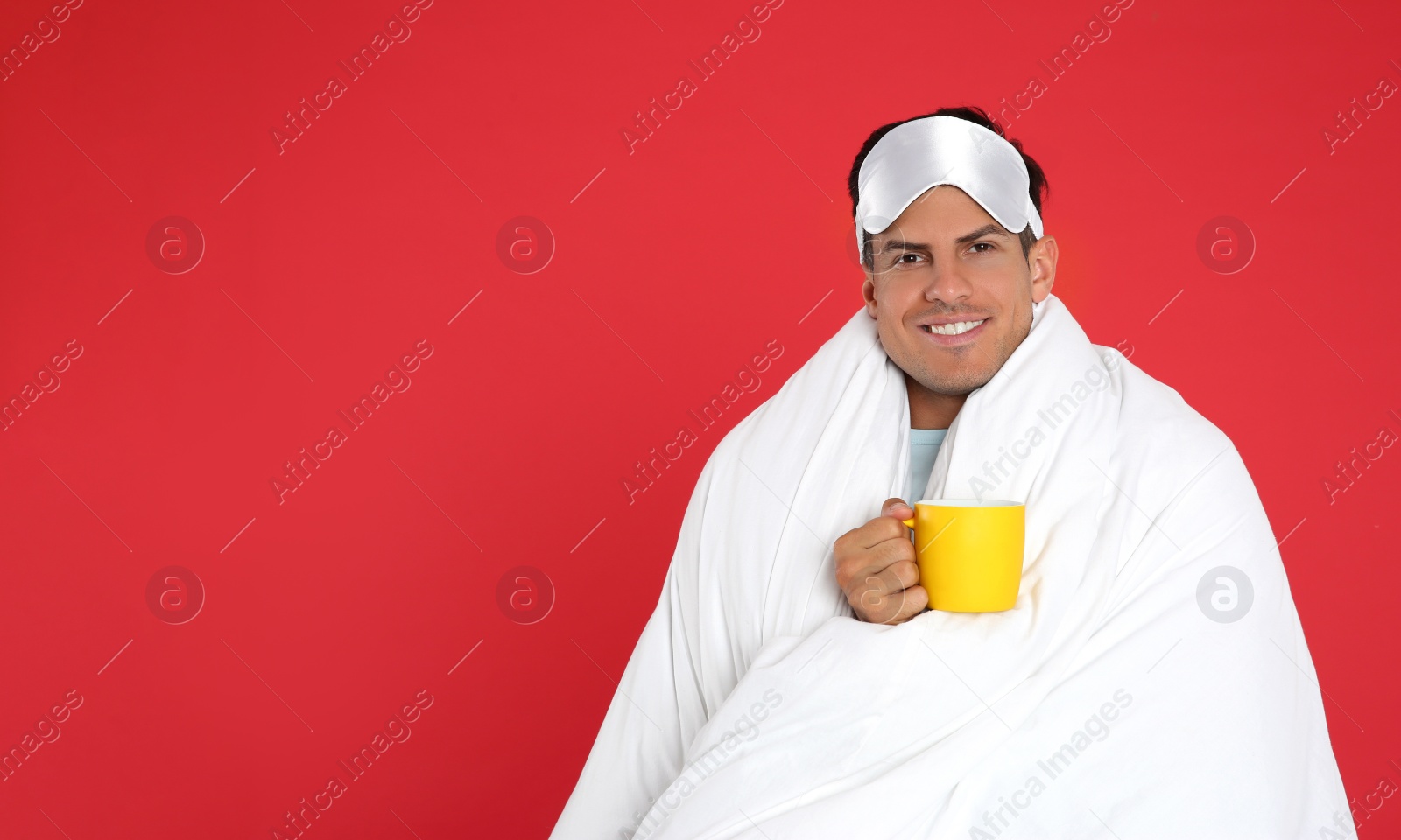 Photo of Man in sleeping mask wrapped with blanket holding cup on red background. Space for text