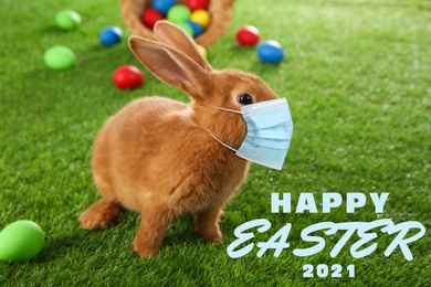 Text Happy Easter 2021 and cute bunny in protective mask on green grass. Holiday during Covid-19 pandemic