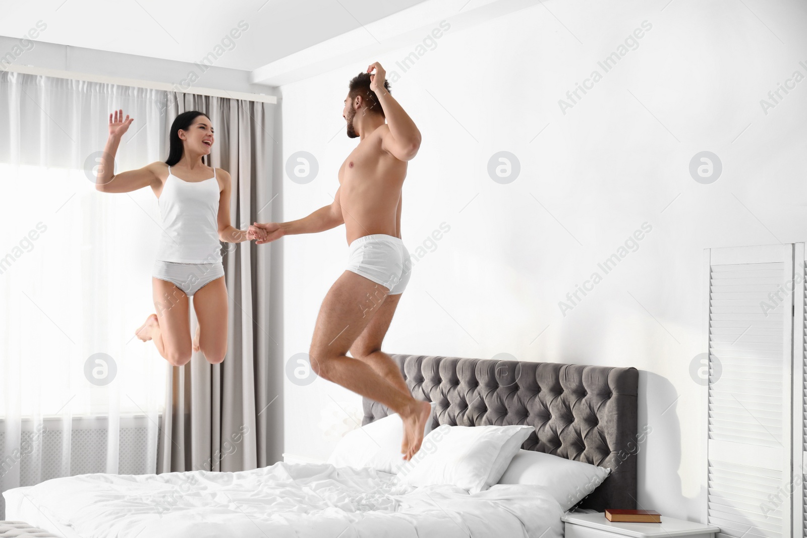 Photo of Young couple in white underwear jumping on bed at home