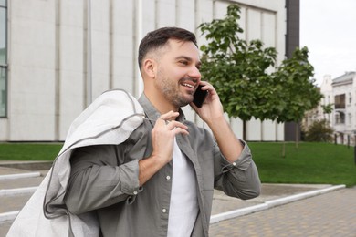 Photo of Man holding garment cover with clothes while talking on phone outdoors. Dry-cleaning service