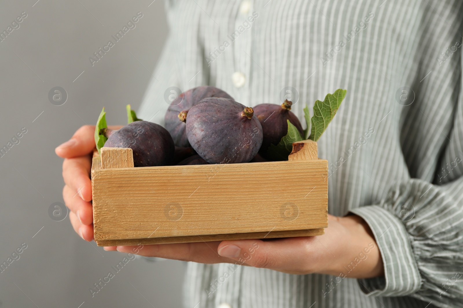 Photo of Woman holding wooden crate with tasty raw figs on light grey background, closeup