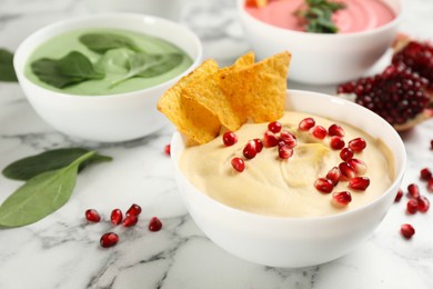 Photo of Bowl of delicious classic hummus with tortilla chips and pomegranate seeds on white marble table