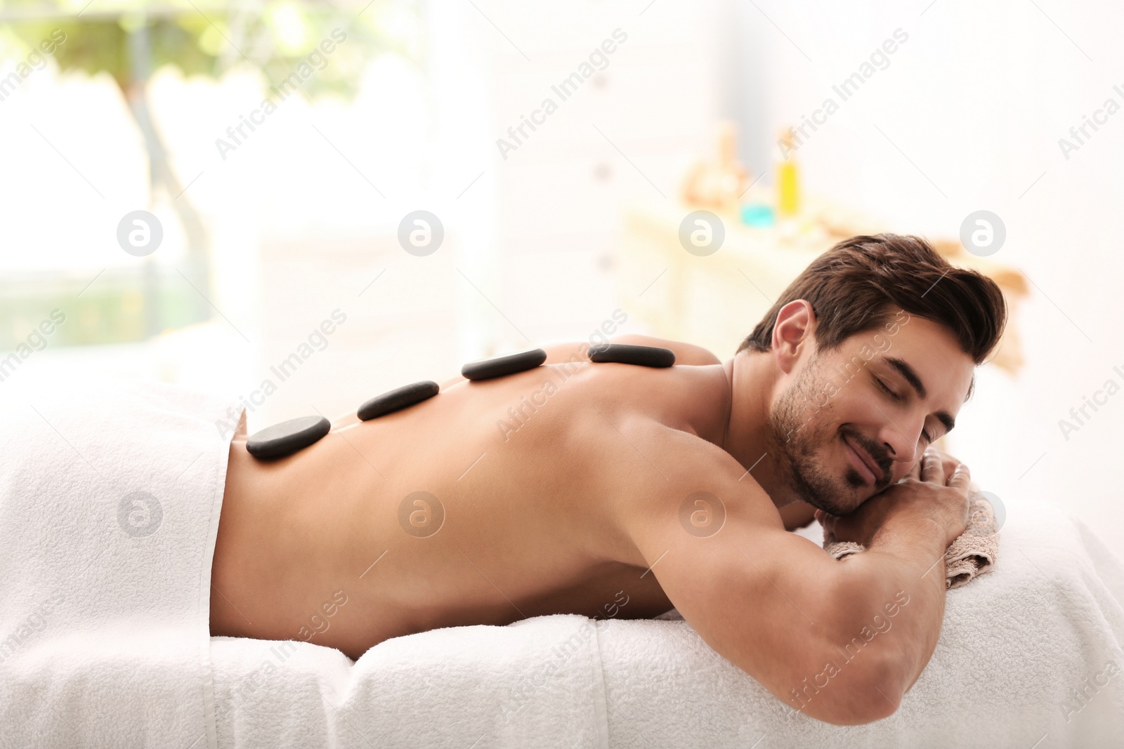 Photo of Handsome young man receiving hot stone massage in spa salon, space for text