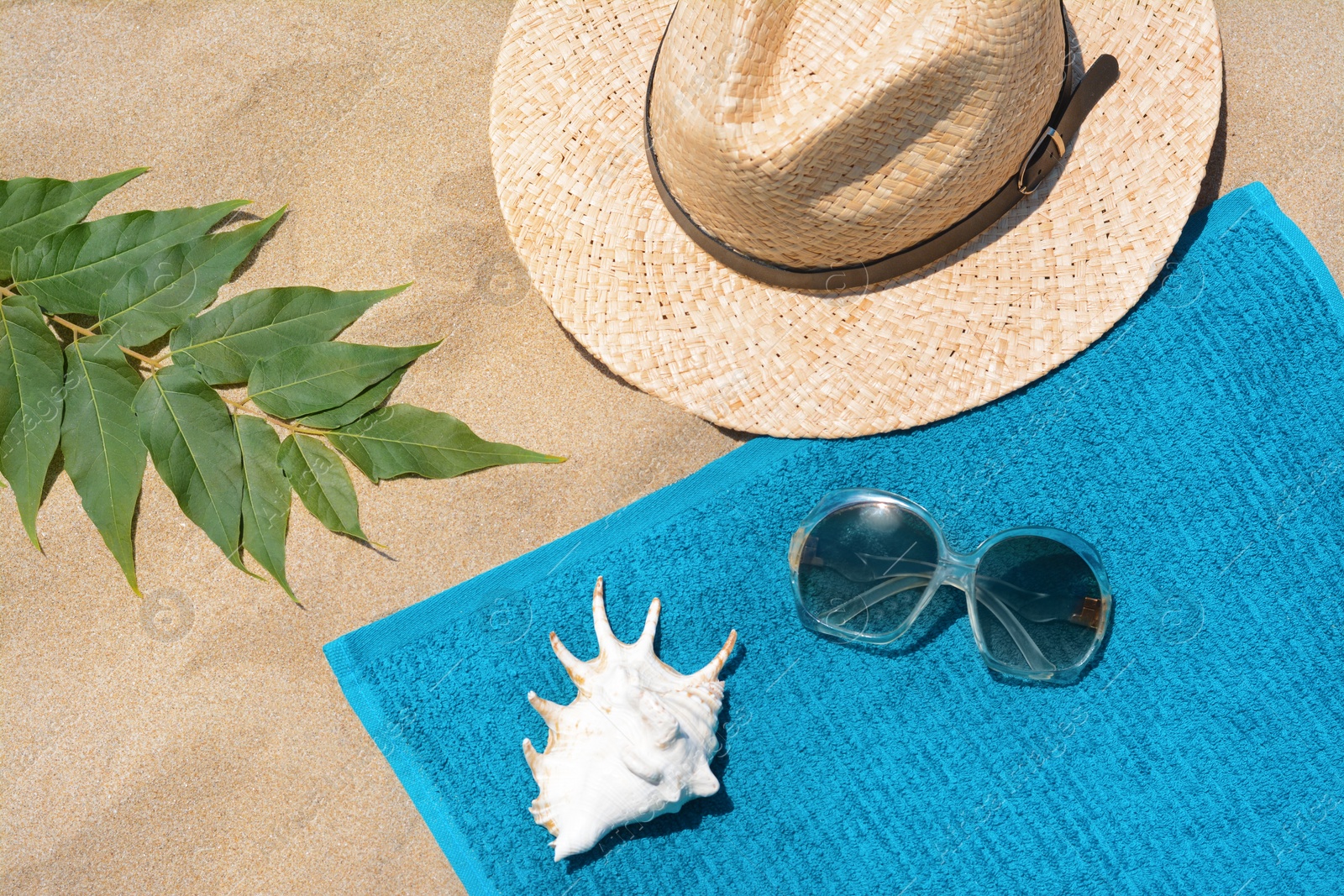 Photo of Soft blue beach towel with sunglasses, straw hat, seashell and green leaves on sand, flat lay