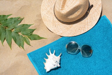 Soft blue beach towel with sunglasses, straw hat, seashell and green leaves on sand, flat lay