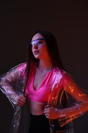 Photo of Fashionable portrait of beautiful woman wearing transparent coat and glasses on dark background in neon lights