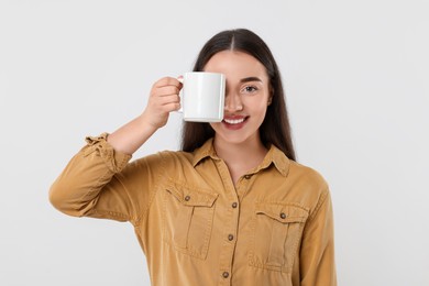 Photo of Happy young woman covering eye with white ceramic mug on light grey background